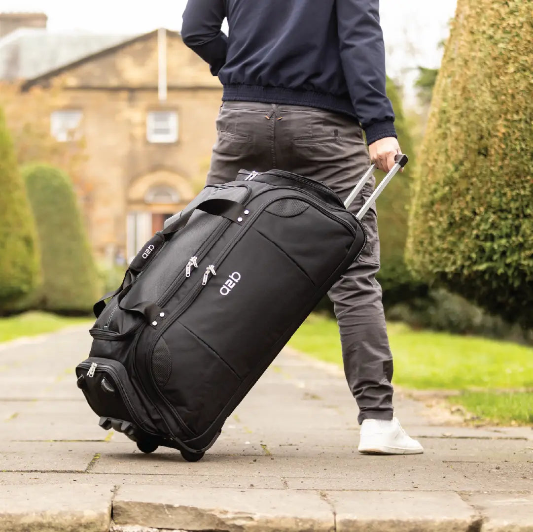 Person pulling a black suitcase with a visible brand logo in an outdoor setting.