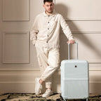 Man in a white outfit holding a light blue suitcase against a neutral wall.