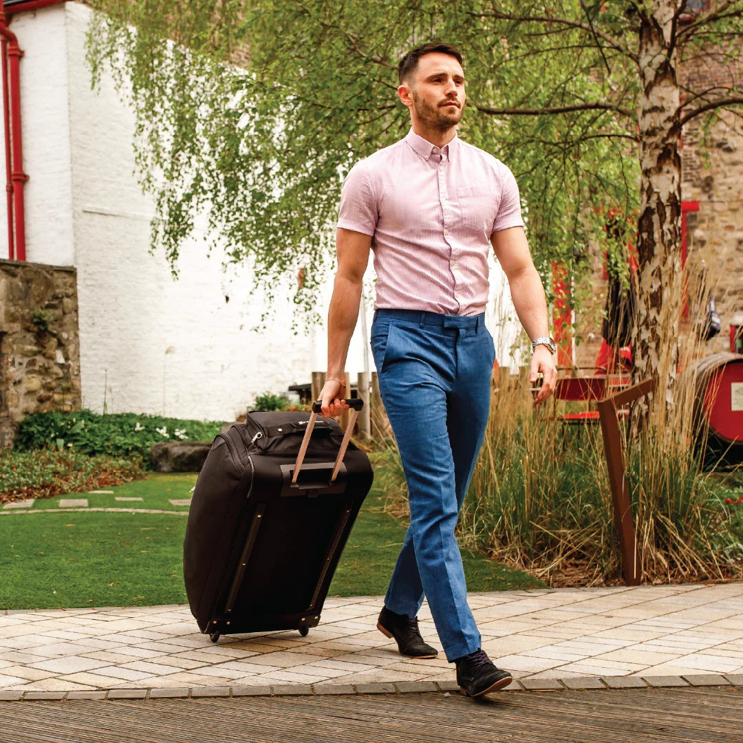 Man in a pink shirt and blue pants pulling a suitcase outdoors.