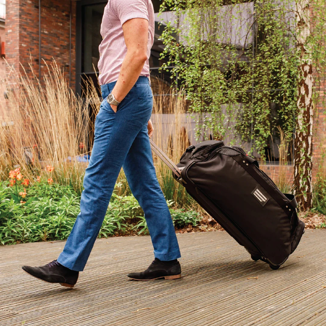 Man pulling a black suitcase outdoors with plants and a building in the background