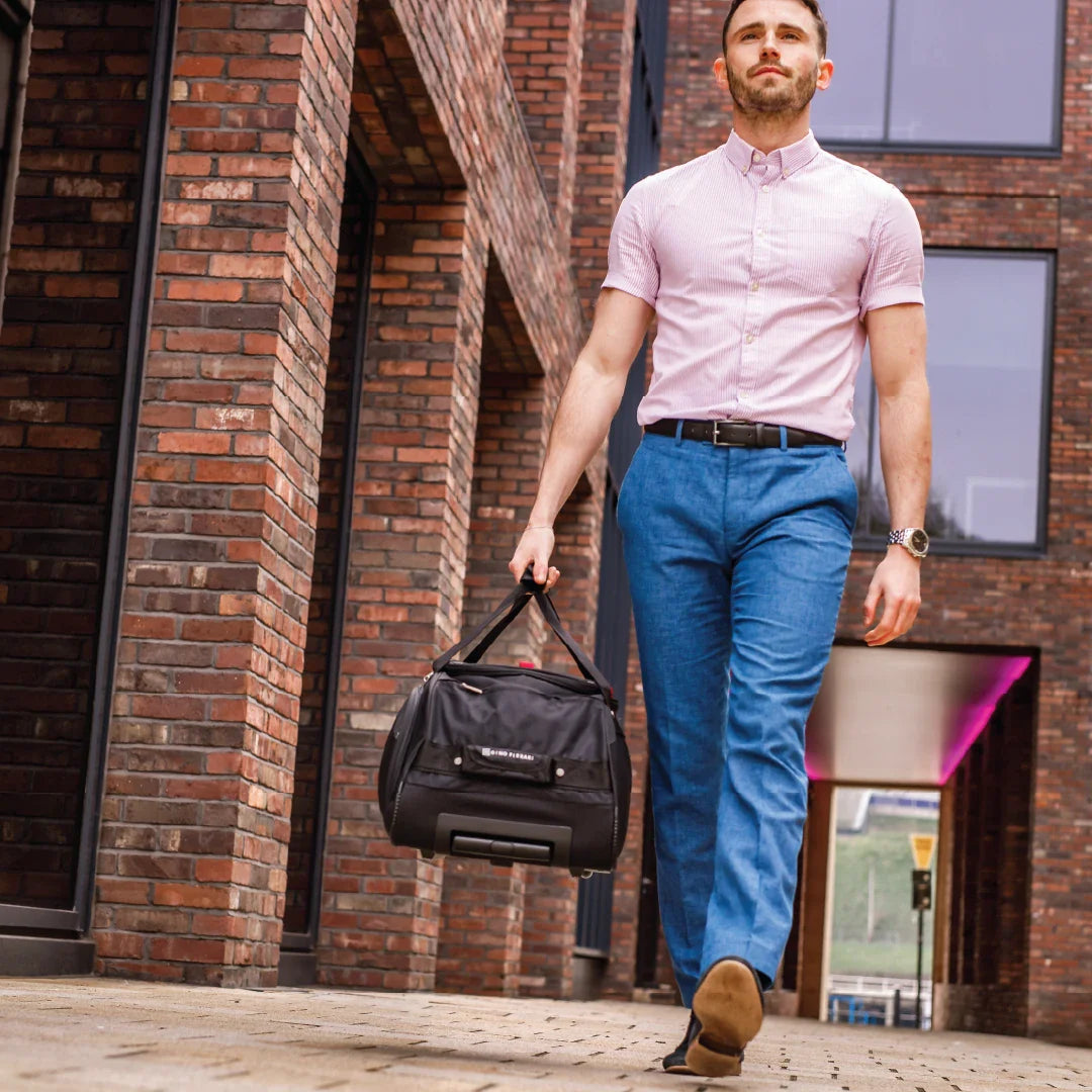 Man in pink shirt and blue pants walking with a black duffel bag against a brick wall.
