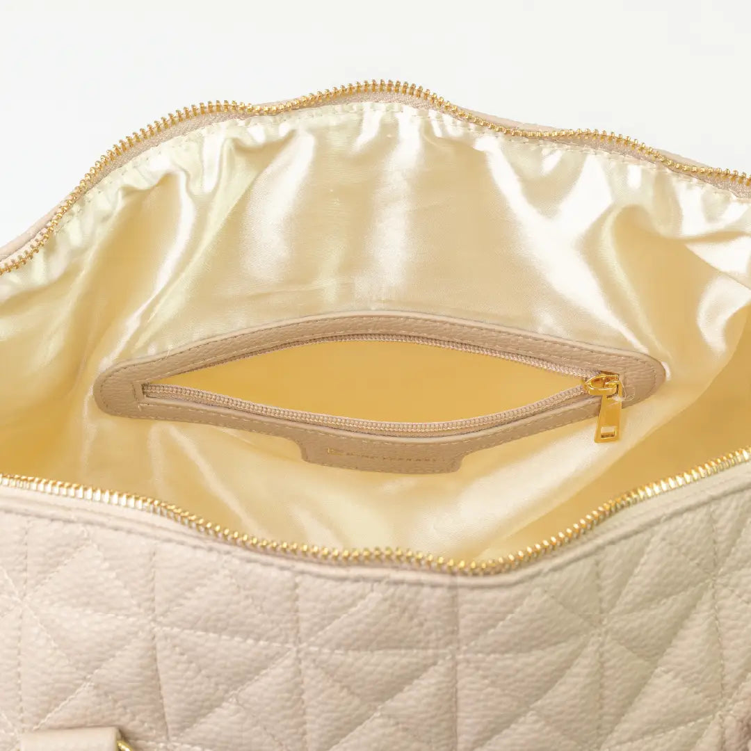 A close-up of a beige bag with cream interior, patterned stitching and a gold zip.