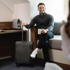 Man sitting in a room with a suitcase and holding a cup, smiling.