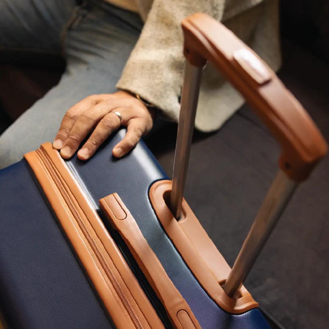 Close-up of a person's hand holding a blue suitcase with brown handle and trim.